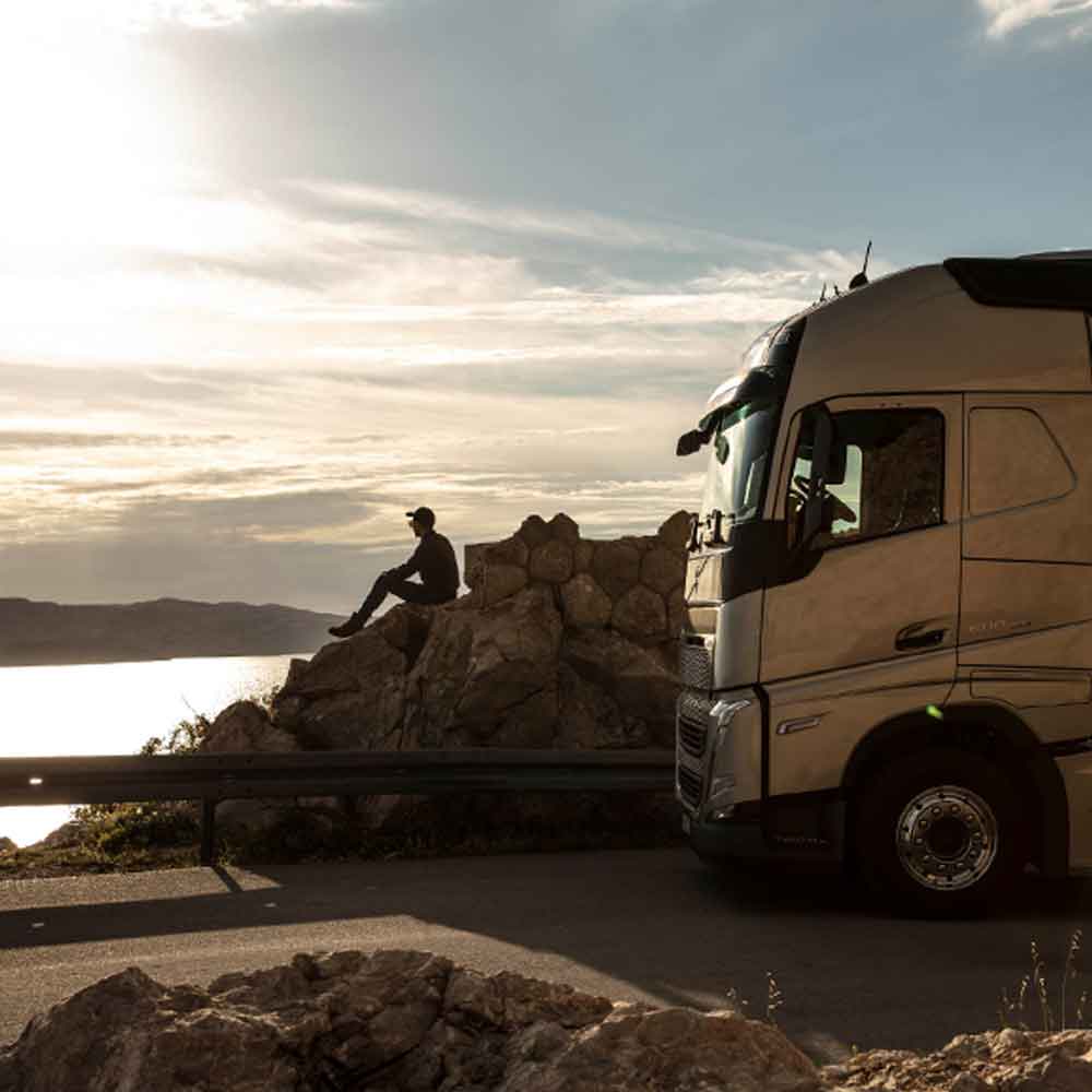 Motorista sentado numa rocha a ver o mar ao pôr-do-sol, com o seu camião Volvo FH estacionado ao seu lado